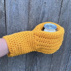Puede incluir: Un portabotellas de ganchillo amarillo brillante hecho a mano, dise&ntilde;ado para mantener las bebidas fr&iacute;as. El portabotellas tiene forma de guante, con un agujero para que la lata se asiente. El portabotellas sostiene una lata plateada. El fondo es una superficie de madera envejecida.