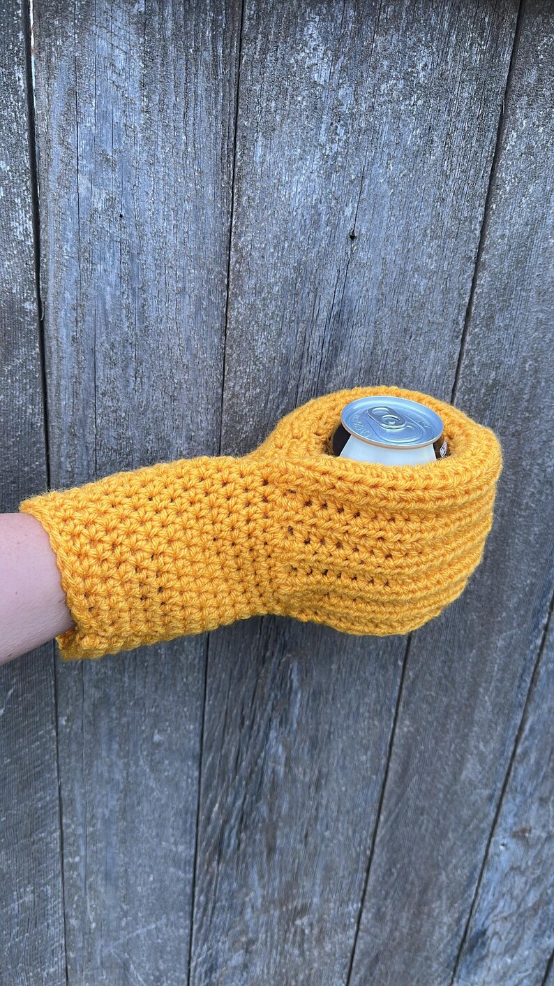 Puede incluir: Un portabotellas de ganchillo amarillo brillante hecho a mano, dise&ntilde;ado para mantener las bebidas fr&iacute;as. El portabotellas tiene forma de guante, con un agujero para que la lata se asiente. El portabotellas sostiene una lata plateada. El fondo es una superficie de madera envejecida.