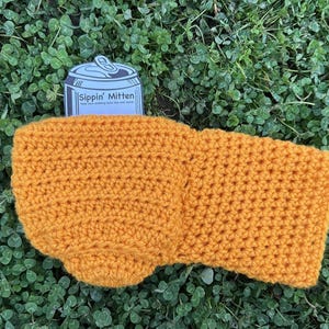 Puede incluir: Funda de bebida de ganchillo amarillo mostaza con una etiqueta en forma de lata que dice "Sippin' Mitten". La funda est&aacute; hecha a mano y descansa sobre un lecho de tr&eacute;bol verde. La funda est&aacute; dise&ntilde;ada para mantener las bebidas fr&iacute;as.