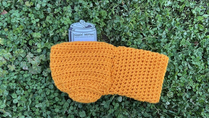 Puede incluir: Funda de bebida de ganchillo amarillo mostaza con una etiqueta en forma de lata que dice "Sippin' Mitten". La funda est&aacute; hecha a mano y descansa sobre un lecho de tr&eacute;bol verde. La funda est&aacute; dise&ntilde;ada para mantener las bebidas fr&iacute;as.