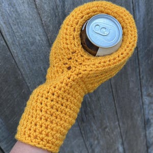 Puede incluir: Un portabotellas de ganchillo amarillo mostaza, dise&ntilde;ado para aislar una lata de bebida de tama&ntilde;o est&aacute;ndar. El portabotellas tiene una manga para el brazo y un ajuste perfecto alrededor de la lata. El fondo es una superficie de madera desgastada.