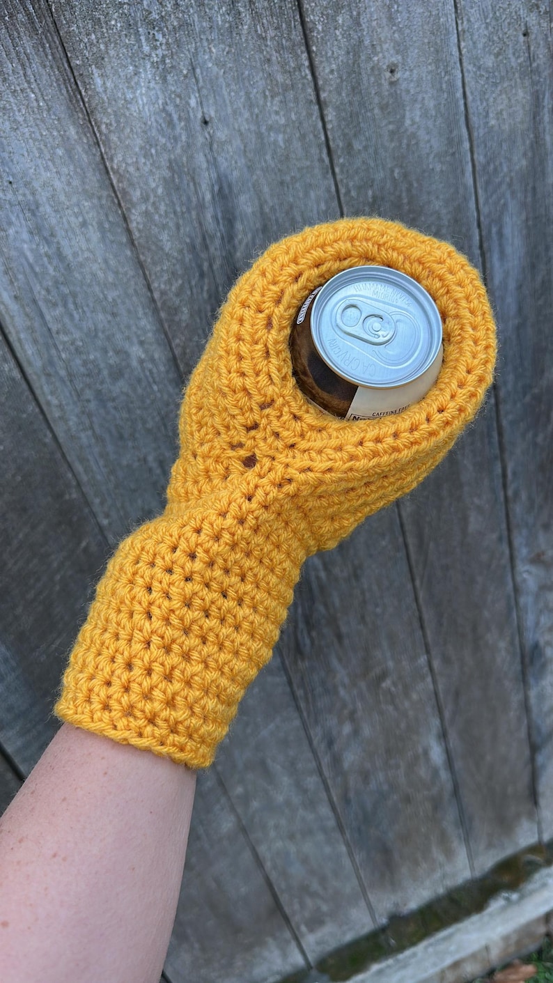 Puede incluir: Un portabotellas de ganchillo amarillo mostaza, dise&ntilde;ado para aislar una lata de bebida de tama&ntilde;o est&aacute;ndar. El portabotellas tiene una manga para el brazo y un ajuste perfecto alrededor de la lata. El fondo es una superficie de madera desgastada.
