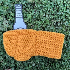 Puede incluir: Un soporte de botella de cerveza tejido a ganchillo de color amarillo mostaza con el texto "Sippin' Mitten" en el abrebotellas. El soporte est&aacute; hecho de un hilo texturizado y est&aacute; dise&ntilde;ado para mantener las bebidas fr&iacute;as. El soporte est&aacute; sobre un lecho de tr&eacute;bol verde.