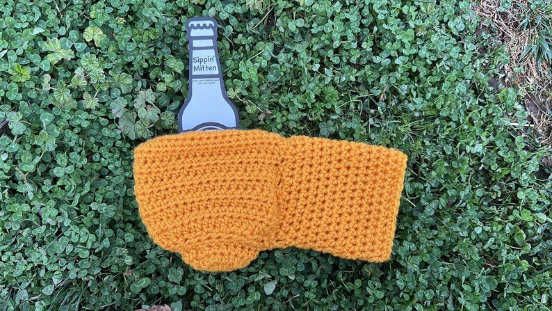 Puede incluir: Un soporte de botella de cerveza tejido a ganchillo de color amarillo mostaza con el texto "Sippin' Mitten" en el abrebotellas. El soporte est&aacute; hecho de un hilo texturizado y est&aacute; dise&ntilde;ado para mantener las bebidas fr&iacute;as. El soporte est&aacute; sobre un lecho de tr&eacute;bol verde.