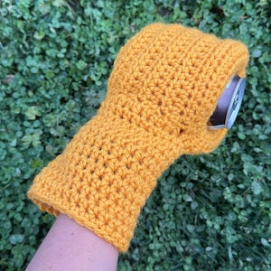 Puede incluir: Funda para bebidas de ganchillo amarillo mostaza. La funda est&aacute; dise&ntilde;ada para adaptarse a una lata est&aacute;ndar y est&aacute; hecha a mano. La funda se muestra en el brazo de una persona, con un fondo verde de tr&eacute;bol.