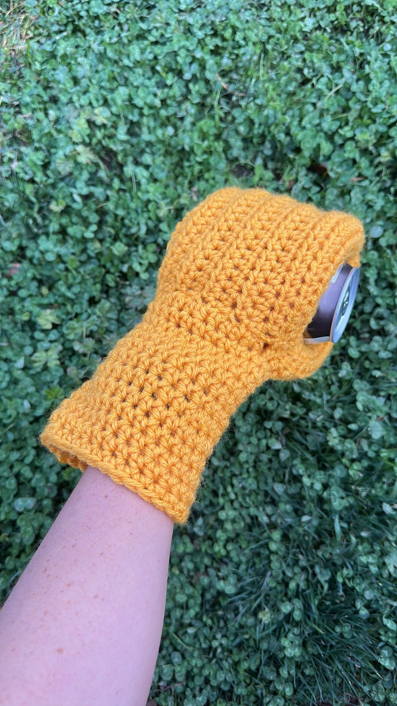 Puede incluir: Funda para bebidas de ganchillo amarillo mostaza. La funda est&aacute; dise&ntilde;ada para adaptarse a una lata est&aacute;ndar y est&aacute; hecha a mano. La funda se muestra en el brazo de una persona, con un fondo verde de tr&eacute;bol.