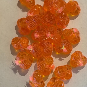 May include: A collection of small, orange, heart-shaped objects encased in orange netting. Each bundle is tied with a small pink accent. The objects are scattered across a light-colored surface, creating a visually appealing arrangement.