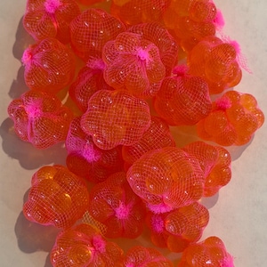 Op de afbeelding: Een verzameling oranje, bloemvormige objecten, elk verpakt in een roze net. De objecten hebben een doorschijnend uiterlijk en het net is aan de bovenkant vastgebonden met een roze touwtje. Het geheel is helder en kleurrijk.