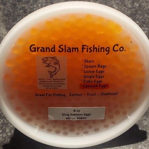 Puede incluir: Un recipiente blanco lleno de huevas de pescado de color naranja brillante, etiquetado como "Grand Slam Fishing Co." con texto que incluye "Chinook Eggs" y "King Salmon Eggs." El recipiente contiene 227g de huevos, ideal para la pesca.