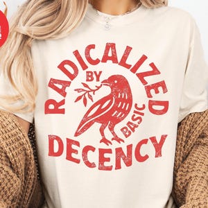 May include: Cream-colored t-shirt with a red graphic that reads "RADICALIZED BY BASIC DECENCY" around a red bird holding a sprig. The shirt is part of a casual clothing collection.