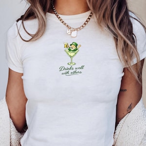 May include: White crop top with a graphic of a leprechaun in a martini glass holding a beer. The text "Drinks well with others" is printed below the graphic in green. The shirt is made of a soft material.