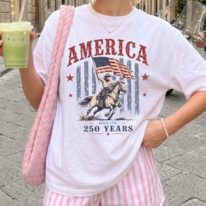 May include: A white short-sleeve t-shirt featuring a graphic print with the text AMERICA, a waving American flag, a cowboy riding a horse, and the text SINCE 1776 250 YEARS. The shirt is paired with pink and white vertical striped shorts.