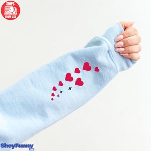 Op de afbeelding: Lichtblauwe sweatshirtmouw met een patroon van rode harten en zwarte sterren. De tekst "SheyFunny" is zichtbaar aan de onderkant. De afbeelding is tegen een witte achtergrond.