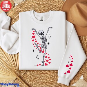 Op de afbeelding: Witte sweatshirt met een dansend skeletontwerp en rode hartaccenten. Het skelet staat in een balletpose, met opgeheven armen en achtervolgende harten. Het ontwerp wordt herhaald op de mouw. Een leuk, grillig ontwerp voor Valentijnsdag of elke dag.