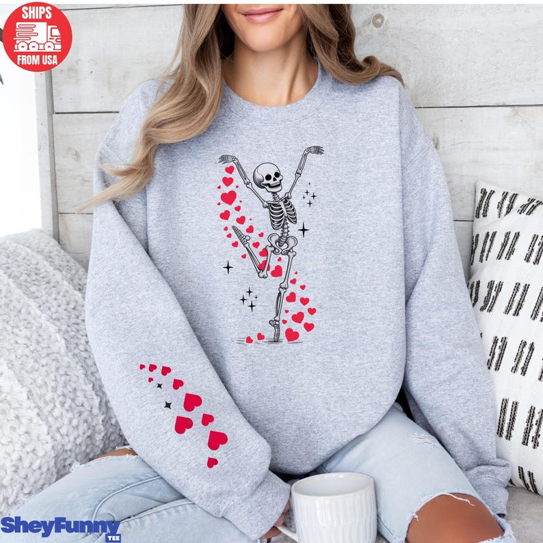 Op de afbeelding: Een lichtgrijs sweatshirt met een dansend skeletontwerp en rode hartaccenten. De linkermouw heeft een patroon van rode harten. Het sweatshirt heeft onderaan de tekst "SheyFunny Tee".