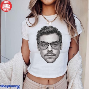 May include: A white short-sleeved t-shirt featuring a large black and white photographic print of a person with a mustache wearing thick-rimmed glasses centered on the chest. The shirt has a crew neckline and is worn with a gold-toned chain necklace.