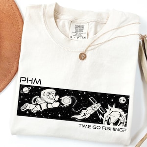 May include: Cream-colored t-shirt with a black and white graphic of an astronaut, a space station, and the text "TIME GO FISHING?". A gold necklace with a pendant is draped on the shirt.