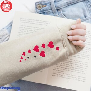 Op de afbeelding: Beige sweatshirtmouw met een ontwerp van rode harten en zwarte sterren. De mouw rust op een open boek. De tekst "SheyFunny Tee" staat onderaan.