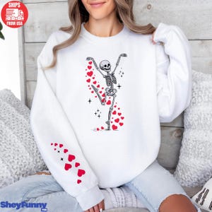 Op de afbeelding: Witte sweatshirt met een dansend skeletontwerp, versierd met rode harten en glitters. Een spoor van rode harten omringt het skelet. De linkermouw heeft een patroon van rode harten. De tekst "SheyFunny Tee" staat onderaan.