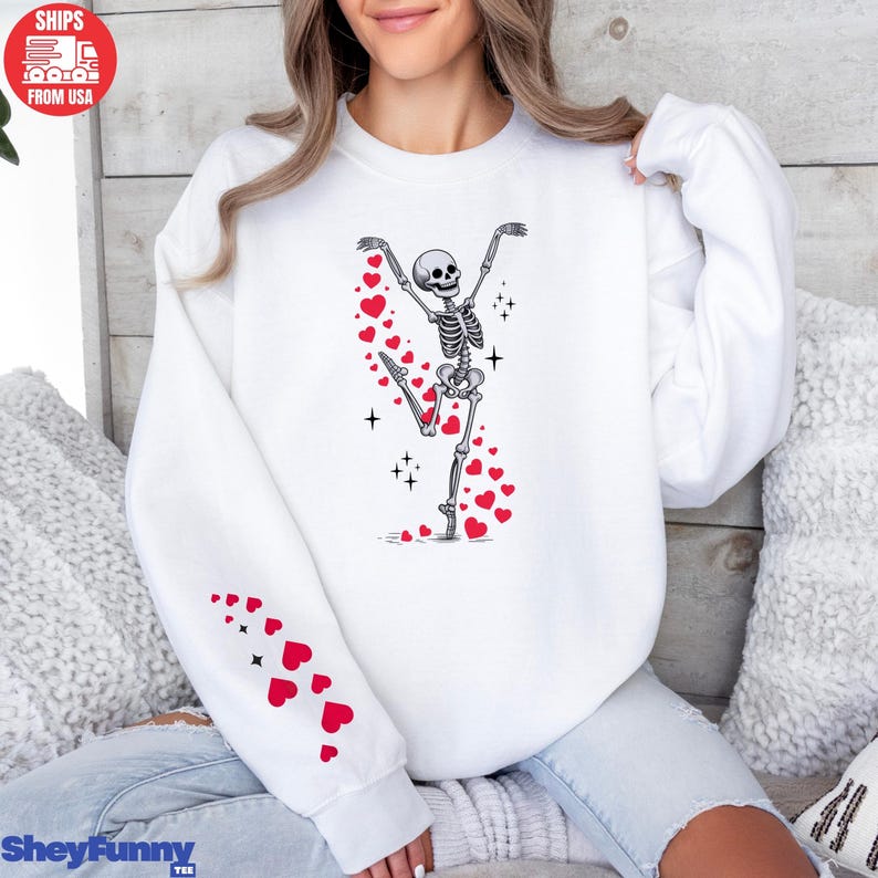Op de afbeelding: Witte sweatshirt met een dansend skeletontwerp, versierd met rode harten en glitters. Een spoor van rode harten omringt het skelet. De linkermouw heeft een patroon van rode harten. De tekst "SheyFunny Tee" staat onderaan.