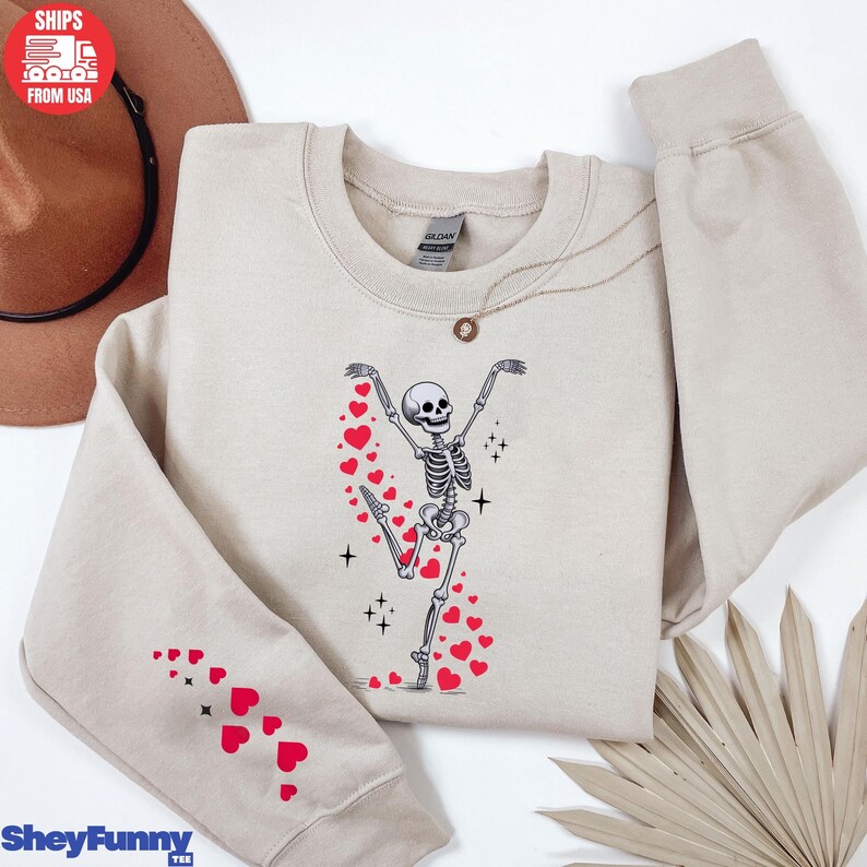 Op de afbeelding: Een beige sweatshirt met een dansende skeletafbeelding met rode harten. De linkermouw heeft een patroon van rode harten. De tekst "SheyFunny Tee" staat linksonder.