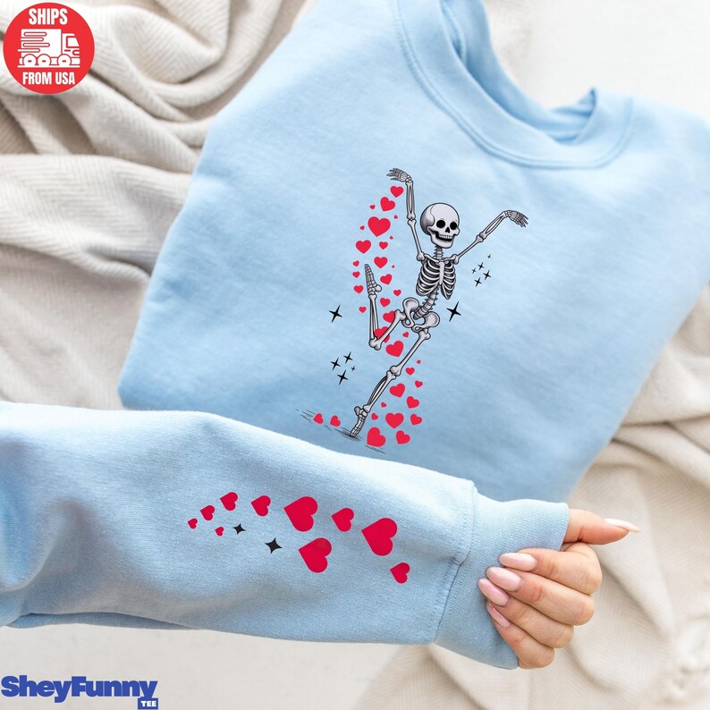 Op de afbeelding: Lichtblauw sweatshirt met een dansend skeletontwerp en rode hartaccenten. Het skelet staat in een balletpose, met harten die erachteraan komen. De mouw heeft ook rode hart- en sterontwerpen. De tekst "SheyFunny" staat onderaan.