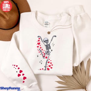 Op de afbeelding: Witte sweatshirt met een dansend skeletontwerp en rode hartaccenten. Het skelet staat in een speelse pose, omringd door harten en sterren. De linkermouw heeft een patroon van rode harten. De sweatshirt heeft de tekst "SheyFunny".
