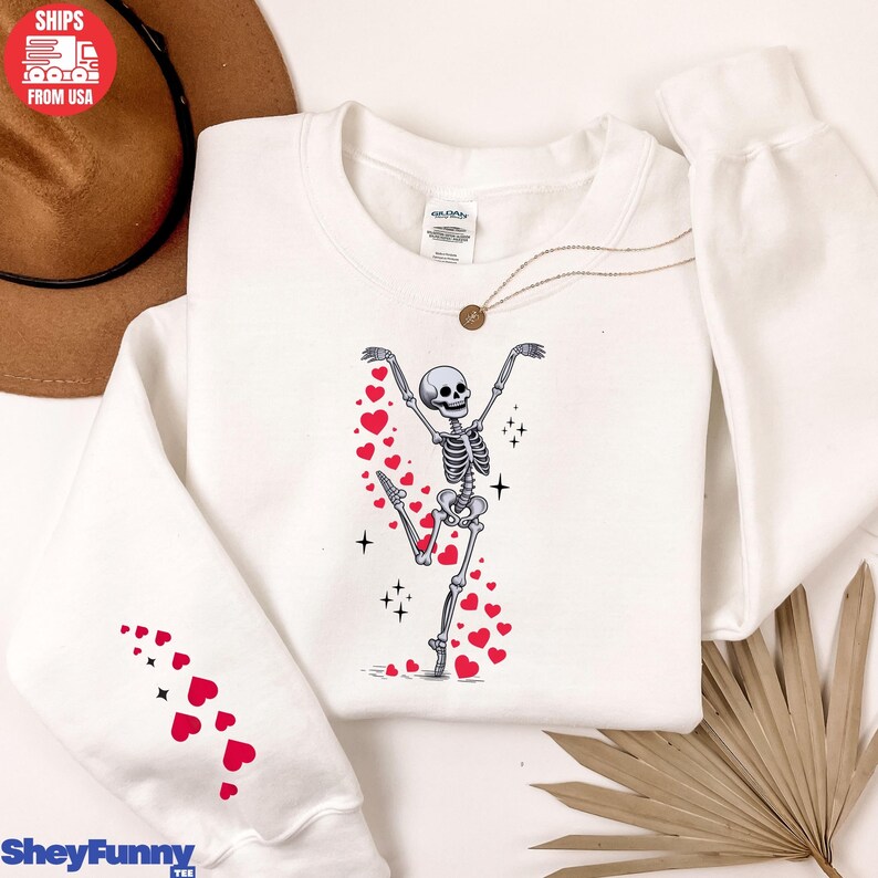 Op de afbeelding: Witte sweatshirt met een dansend skeletontwerp en rode hartaccenten. Het skelet staat in een speelse pose, omringd door harten en sterren. De linkermouw heeft een patroon van rode harten. De sweatshirt heeft de tekst "SheyFunny".