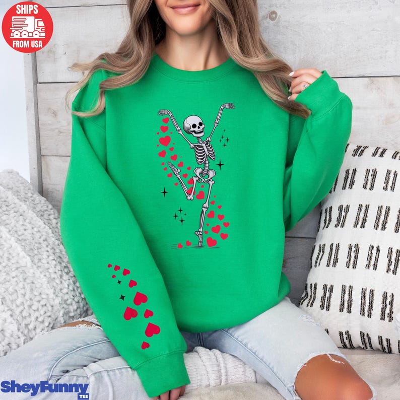 Op de afbeelding: Een felgroene sweatshirt met een dansend skeletontwerp en rode hartaccenten. Het skelet heeft de armen omhoog, en harten komen uit zijn handen en been. De sweatshirt heeft een ronde hals en lange mouwen.