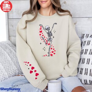 Op de afbeelding: Beige sweatshirt met een dansend skeletontwerp en rode hartdetails. Het skelet staat in een balletpose, met een spoor van harten. Rode harten staan ook op de linkermouw. De tekst "SheyFunny Tee" staat onderaan.