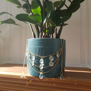 Puede incluir: Una maceta de cerámica turquesa con una cadena dorada y adornos de cristal. La maceta contiene una planta verde. La cadena dorada tiene múltiples hebras con colgantes de cristal transparente y cuentas de cristal facetadas.