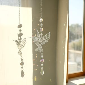 May include: Two hummingbird suncatchers hanging from chains. One is a silhouette with dark accents, the other is a detailed white design with iridescent crystals. The suncatchers are near a window, catching the light.