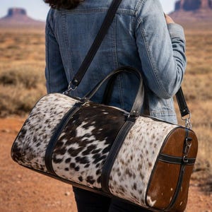 May include: A cowhide duffel bag with a spotted pattern in black and white, and brown leather accents. The bag has a cylindrical shape, black leather straps, and a shoulder strap. The bag is being carried outdoors.