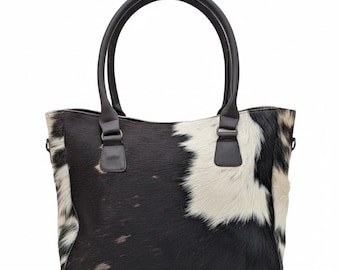 May include: A black and white cowhide tote bag with dark brown leather handles and accents. The bag has a rectangular shape with two rounded handles and a spacious interior. The cowhide pattern is a mix of black and white patches.