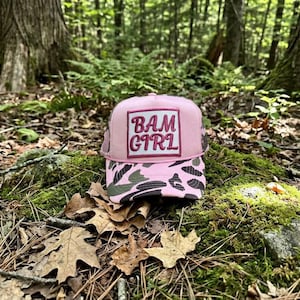 May include: Pink trucker hat with a camouflage brim and the text "BAM GIRL" in a square patch. The hat is set against a backdrop of a forest floor with moss, leaves, and trees.