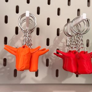 Könnte beinhalten: Schlüsselanhänger mit Miniaturskulpturen der Christusstatue in verschiedenen Farben. Die Schlüsselanhänger sind an silbernen Schlüsselringen und Ketten befestigt. Zu den Farben gehören Beige, Orange, Rot und Grün. Die Schlüsselanhänger werden auf einer weißen Lochplatte ausgestellt.