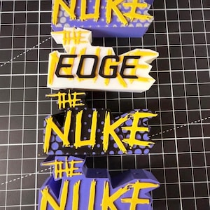 Könnte beinhalten: Vier 3D-gedruckte Schilder mit den Wörtern "THE NUKE" und "THE EDGE". Die Schilder sind in Lila, Weiß und Schwarz gehalten, mit gelben Buchstaben. Der Hintergrund ist ein schwarzes Gitter. Die Schilder scheinen aus Kunststoff zu sein.