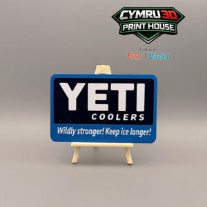 May include: A rectangular blue and black sign featuring the white text YETI COOLERS and the slogan Wildly stronger! Keep ice longer! The sign is displayed on a small wooden easel. Above the sign is a logo for CYMRU 3D PRINT HOUSE.