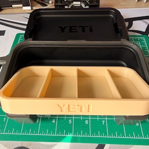 May include: Black YETI-branded storage box with a tan interior tray divided into four compartments. The box is open, revealing the interior. The box is on a green and gray cutting mat.