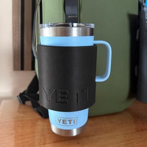 以下が含まれることがあります： ハンドルと透明な蓋が付いたライトブルーのステンレス製タンブラー。YETIのロゴがエンボス加工された黒いスリーブに収められています。ライトブルーのタンブラー本体の下部には、小さな白いYETIのロゴがプリントされています。