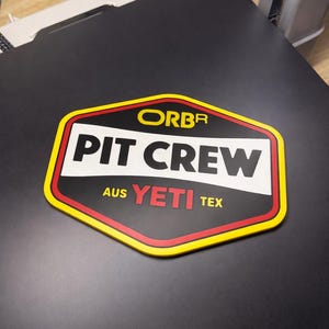 May include: A black, red, white, and yellow Pit Crew sticker. The sticker has the words "ORBR PIT CREW AUS YETI TEX" in white and red lettering. The sticker is on a black surface.