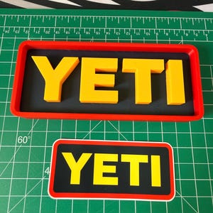 Könnte beinhalten: Zwei rechteckige Objekte mit dem Wort "YETI" in gelben Blockbuchstaben. Eines ist ein 3D-gedrucktes Schild mit rotem Rand und schwarzem Hintergrund. Das andere ist ein Aufkleber mit schwarzem Hintergrund und weißem Rand.