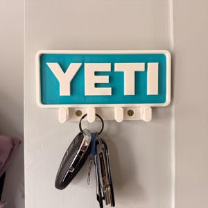 Könnte beinhalten: Ein weißer und türkisfarbener Schlüsselhalter mit dem Wort "YETI" in fetten weißen Buchstaben. Der Schlüsselhalter hat drei Haken und ist an einer hellgrauen Tür montiert. Ein Satz Schlüssel hängt an den Haken.
