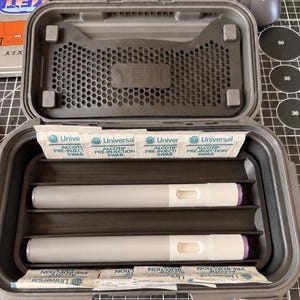 May include: An open, gray YETI case with internal compartments holding two medical devices and several packets of pre-injection swabs. The case is on a black surface with a computer mouse and target stickers visible.