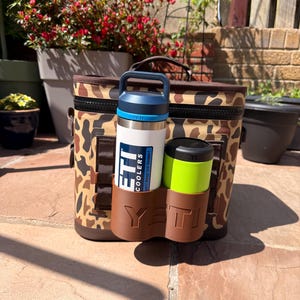 May include: A camouflage-patterned cooler bag with a black zipper and handle. A white and blue water bottle and a green and black tumbler are held in a brown holder attached to the cooler. The cooler and accessories are branded with the "YETI" logo.