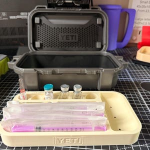 May include: An open gray YETI case with a beige tray containing syringes and vials. The tray has the YETI logo. The case is on a black and white grid surface. A purple mug is in the background.