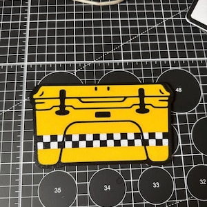 K&ouml;nnte beinhalten: Ein gelber Aufkleber in Form eines Taxi. Der Aufkleber hat schwarze Details, einschlie&szlig;lich der Umrisse, Griffe und ein Schachbrettmuster am unteren Rand. Der Aufkleber befindet sich auf einer schwarzen Schneidematte mit wei&szlig;en Rasterlinien und kreisf&ouml;rmigen Aussparungen.