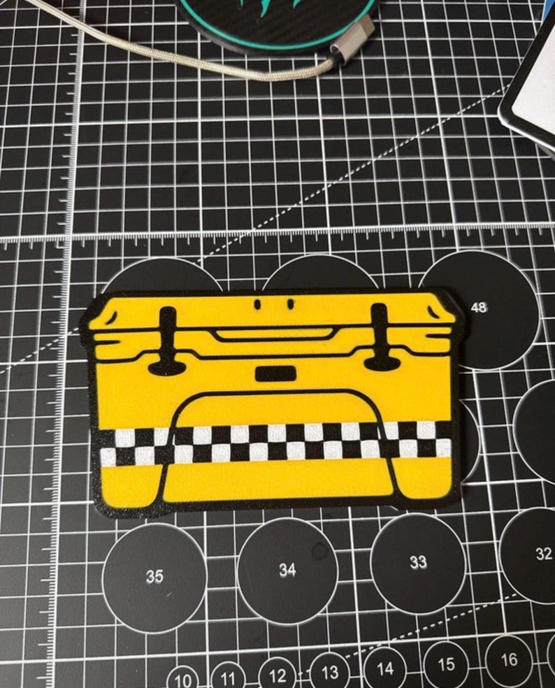 K&ouml;nnte beinhalten: Ein gelber Aufkleber in Form eines Taxi. Der Aufkleber hat schwarze Details, einschlie&szlig;lich der Umrisse, Griffe und ein Schachbrettmuster am unteren Rand. Der Aufkleber befindet sich auf einer schwarzen Schneidematte mit wei&szlig;en Rasterlinien und kreisf&ouml;rmigen Aussparungen.