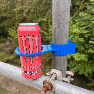 Peut inclure: Une canette de boisson énergisante Monster rouge et noire dans un support bleu fixé à un poteau métallique. La canette porte les mots "Pipeline Punch" et "Punch Monster". Le support est imprimé en 3D et conçu pour une canette standard.