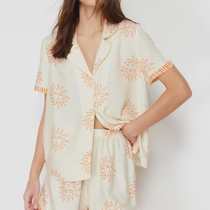 Op de afbeelding: Cr&egrave;mekleurige pyjamaset met een herhalend oranje zonontwerp. De set bevat een overhemd met korte mouwen en bijpassende shorts. Het overhemd heeft een reverskraag en de shorts hebben een elastische tailleband.